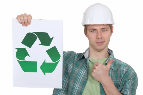 A young man wearing a white safety helmet and a green checked shirt is standing outdoors in front of a plain background, holding a large white sign featuring a green recycling symbol composed of three chasing arrows. His expression is neutral, and with his right hand, he is giving a thumbs-up gesture while his left hand extends to hold the sign up. The setting appears to be a professional environment, possibly related to waste management or recycling services, fitting within the context of rubbish removal. The prominent recycling symbol emphasizes environmental responsibility, which aligns with the services offered by Business Waste Removal Notting Hill. The image displays clear, high-quality details, with textures of the helmet and shirt visible, and the focus is on the individual and the recycling sign. The overall scene underscores themes of sustainability and waste management, relevant to the company's offerings in the Notting Hill area, postcode inferred from the page URL.