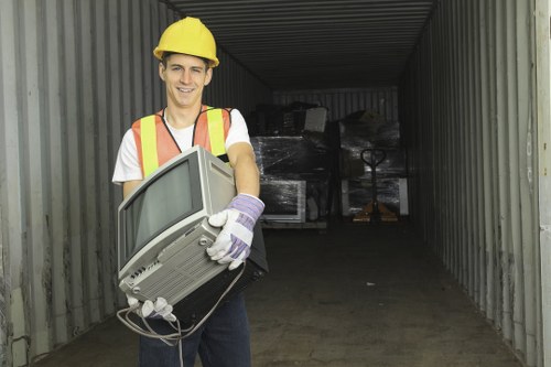 A young male worker wearing a yellow safety helmet, high-visibility vest, and white gloves is standing inside a shipping container, holding an old desktop computer with a CRT monitor, keyboard, and mouse. The computer is beige with a textured plastic exterior, showing signs of age and use. The container's interior has corrugated metal walls with a dull gray finish, and the floor is wooden. In the background, stacked black plastic or cardboard boxes are visible, with some wrapping or packaging material on top. The scene is lit by natural light entering from the open end of the container, creating a well-illuminated environment suitable for specialist rubbish removal services in the Notting Hill area, London. Business Waste Removal Notting Hill provides efficient waste clearance, including old electronics and industrial materials, within the local postcode region.