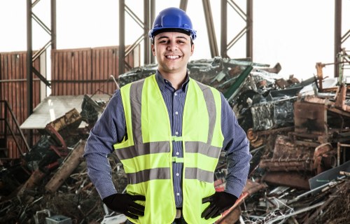 A male waste management worker wearing a blue safety helmet, a high-visibility yellow vest, and black gloves is standing outdoors inside a large industrial warehouse or storage facility. Behind him, there is a pile of assorted rubbish, including damaged metal parts, wood, and plastic waste, which appears to be debris from demolition or large-scale clearance. The background features metal framework and windows that allow natural light to illuminate the scene. The worker is smiling, with his hands resting on his hips, conveying a professional and approachable demeanor. The setting suggests a professional rubbish removal context, consistent with services provided by Business Waste Removal Notting Hill, servicing areas around London with waste disposal needs. The environment is industrial, and the overall scene emphasizes safety, cleanup, and waste management activities suitable for a commercial or construction site in or near Notting Hill.