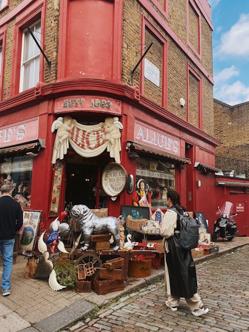 A busy street scene in Portobello Road showing pedestrians walking along the pavement and a row of parked cars lining the curb on the left side. The buildings on the left are multi-storey commercial s